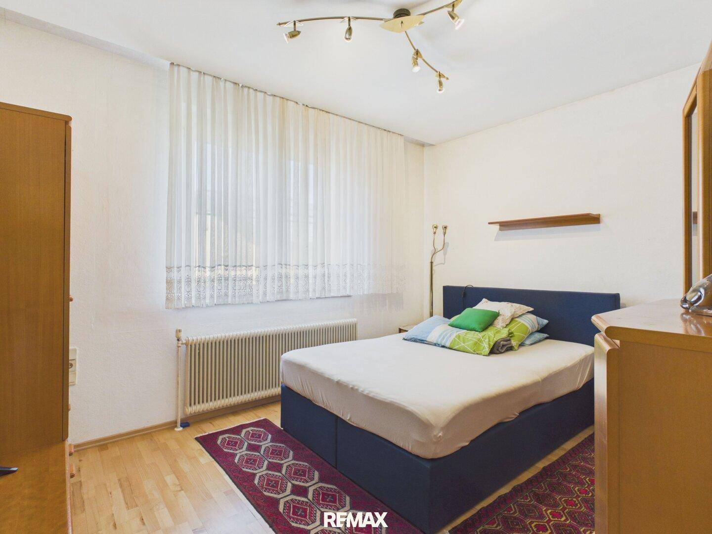 Schlafzimmer mit blauem Bett, Holzfußboden und großem Fenster mit Vorhängen.