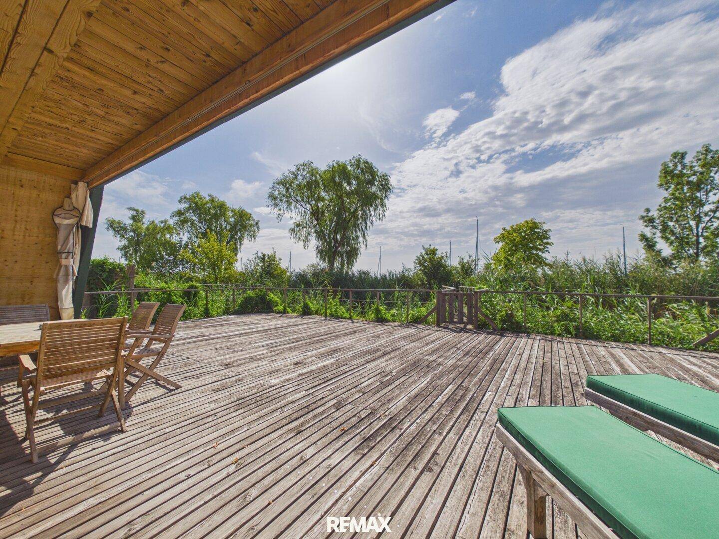 Geräumige Holzterrasse mit Sonnenliegen und Esstisch, umgeben von üppiger Natur.