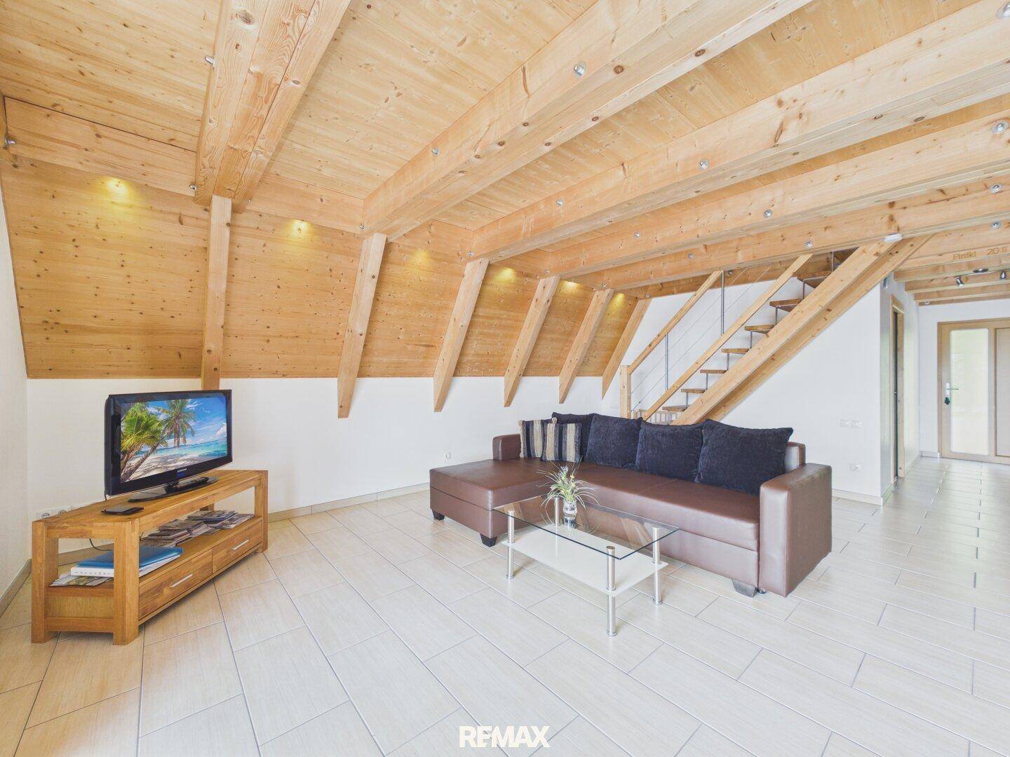 Gemütlicher Wohnbereich mit Holzbalkendecke, Fliesenboden und offener Treppe zum Obergeschoss.