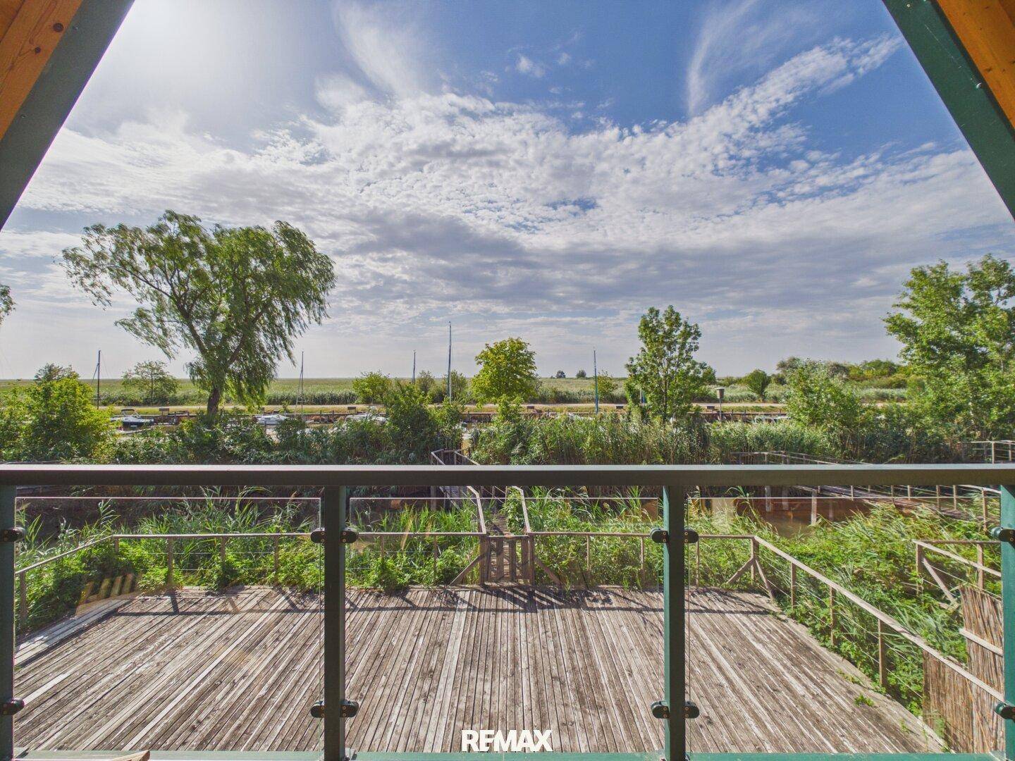 Blick vom Balkon auf die Holzterrasse und die grüne Umgebung unter blauem Himmel.