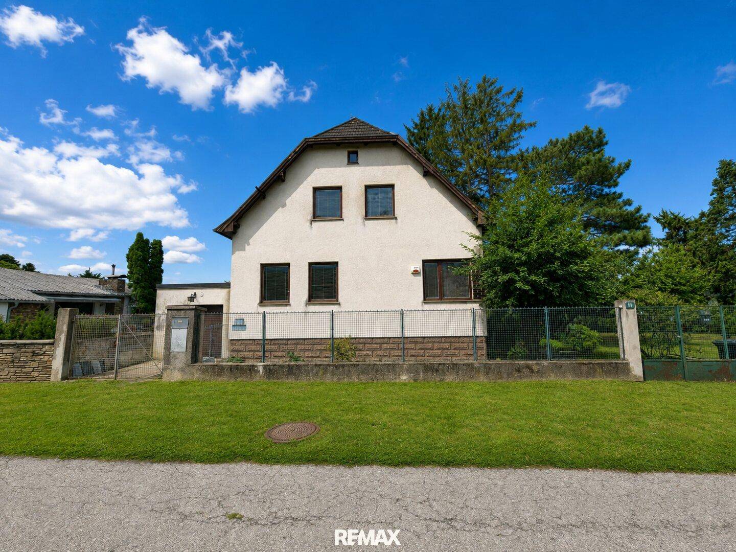Ansicht des Hauses von der Straßenseite mit Zaun, Vorgarten und gepflegtem Rasen unter blauem Himmel.