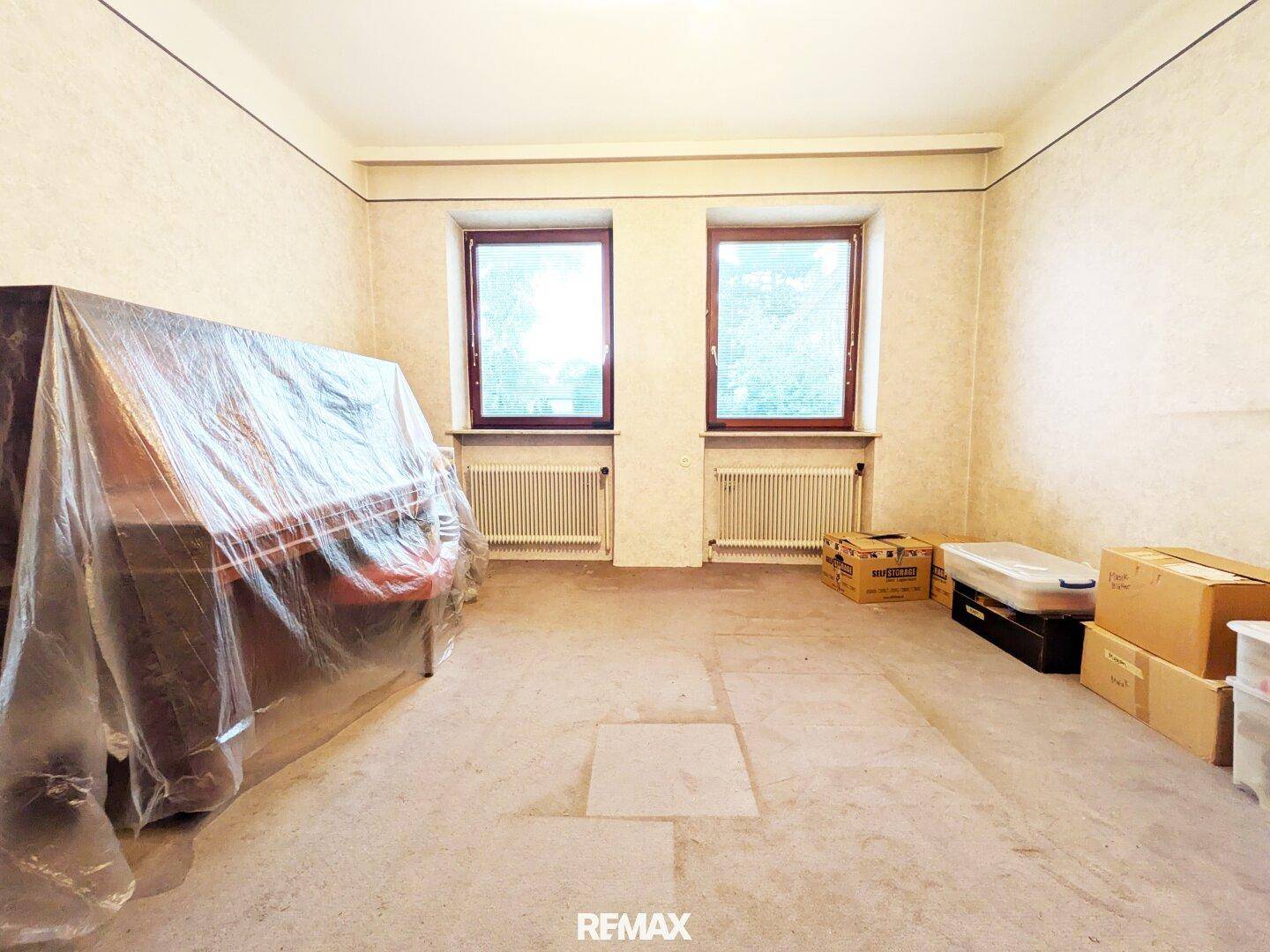 Geräumiger Innenraum mit alter Tapete, zwei Fenstern und einem abgedeckten Klavier, bereit zur Renovierung.