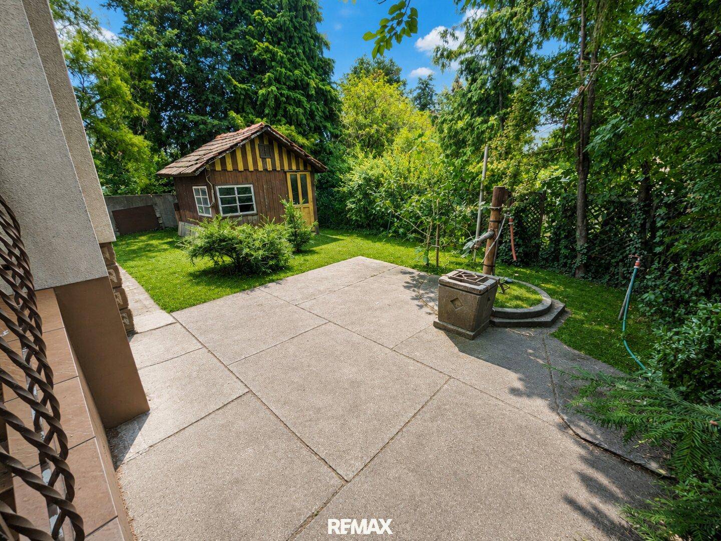 Gepflasterte Terrasse mit Gartenhaus und einem dekorativen Brunnen, umgeben von üppigem Grün.
