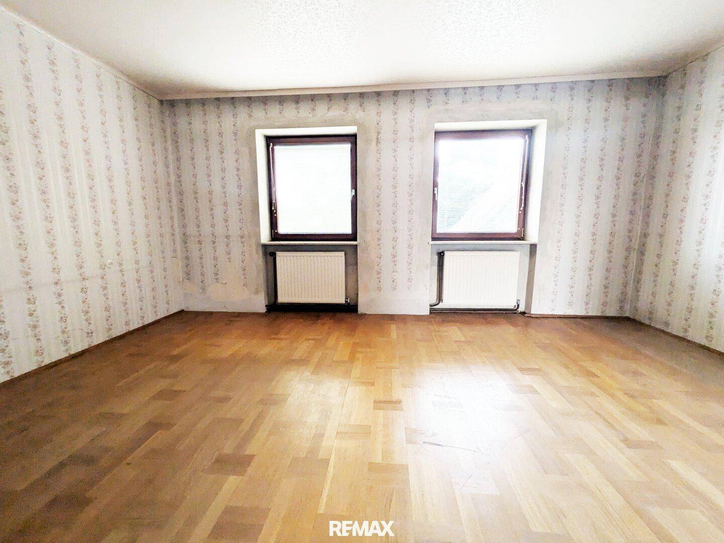 Helles Zimmer mit Holzfußboden, gemusterter Tapete und zwei Fenstern, die den Raum erhellen.
