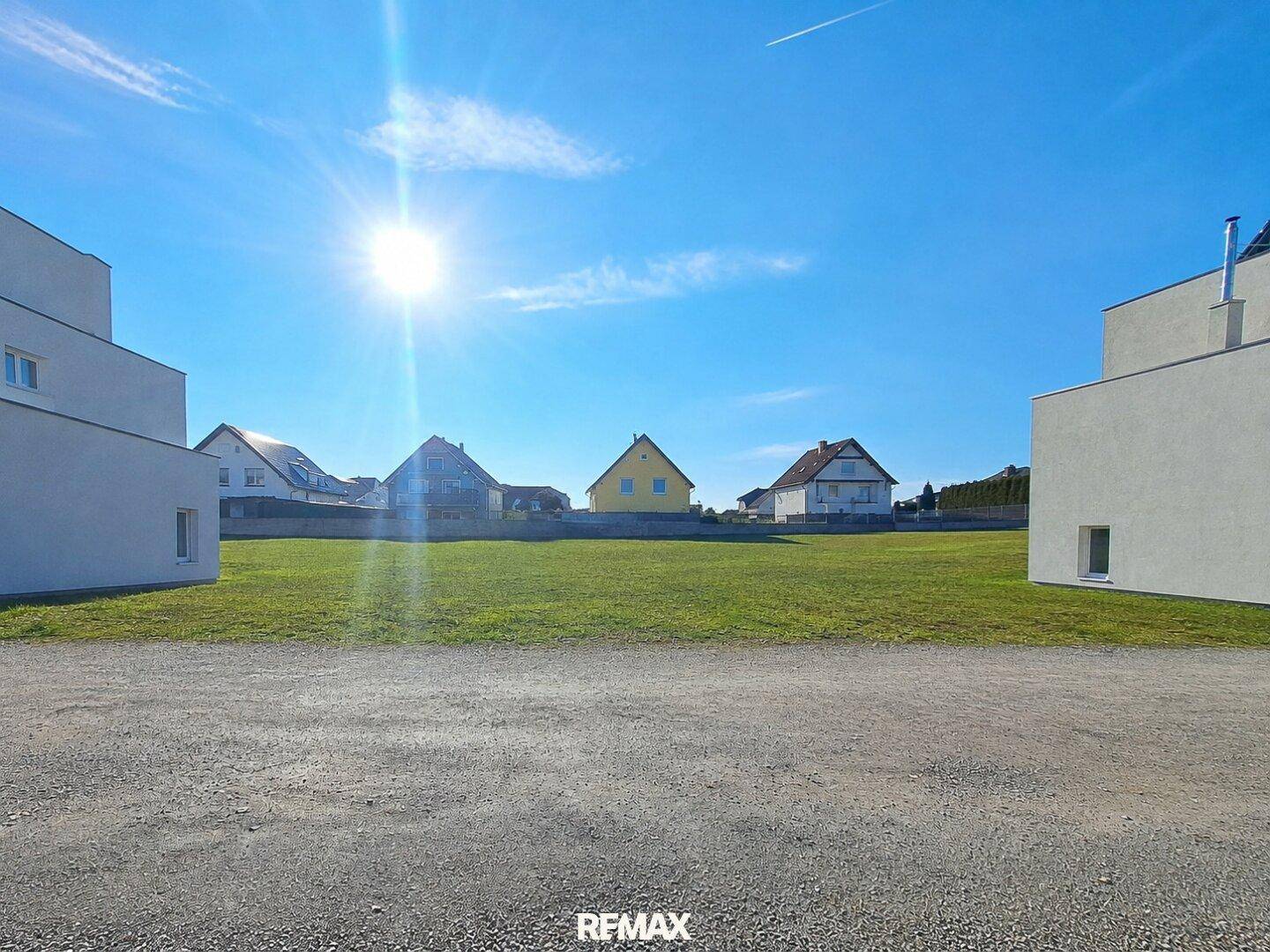 Sonniges Grundstück mit Schotterweg und modernen Gebäuden im Vordergrund, weitere Häuser im Hintergrund.