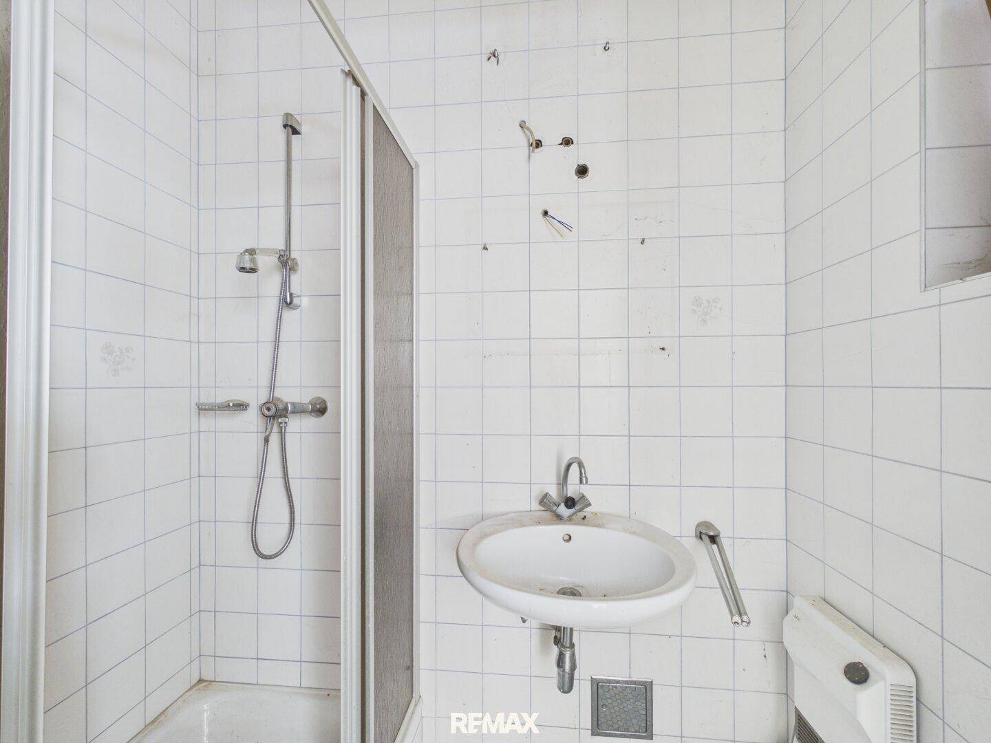 Einfaches Badezimmer mit Duschkabine, Waschbecken und Wandfliesen, sanierungsbedürftig.