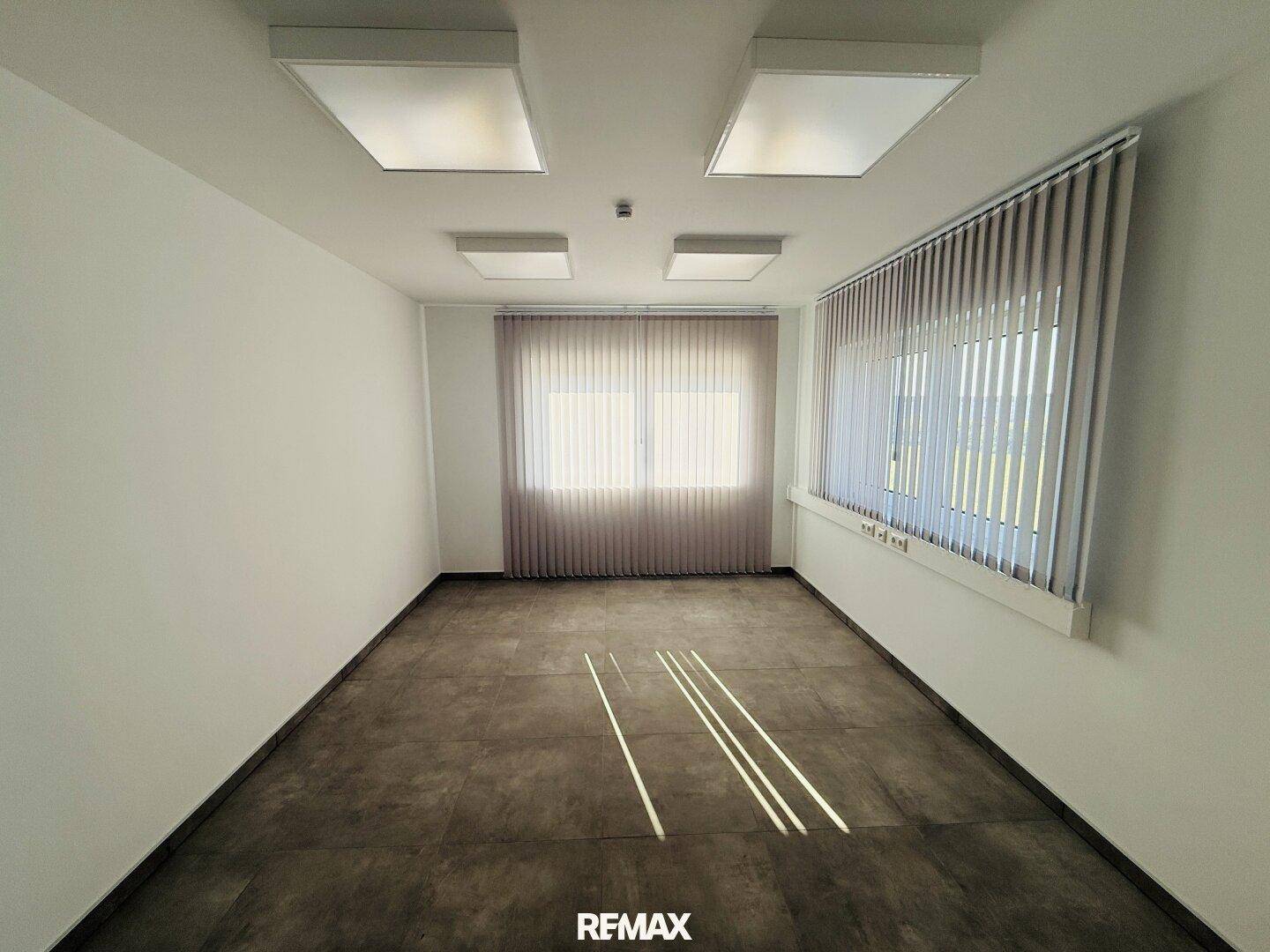 Heller, leerer Raum mit großen Fenstern, Jalousien und modernen Deckenleuchten, ideal als Büro.