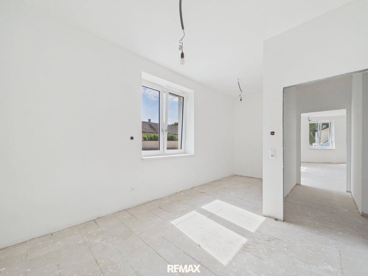 Geräumiges Zimmer im Rohbau mit Fenster und offener Verbindung zu einem weiteren Raum.