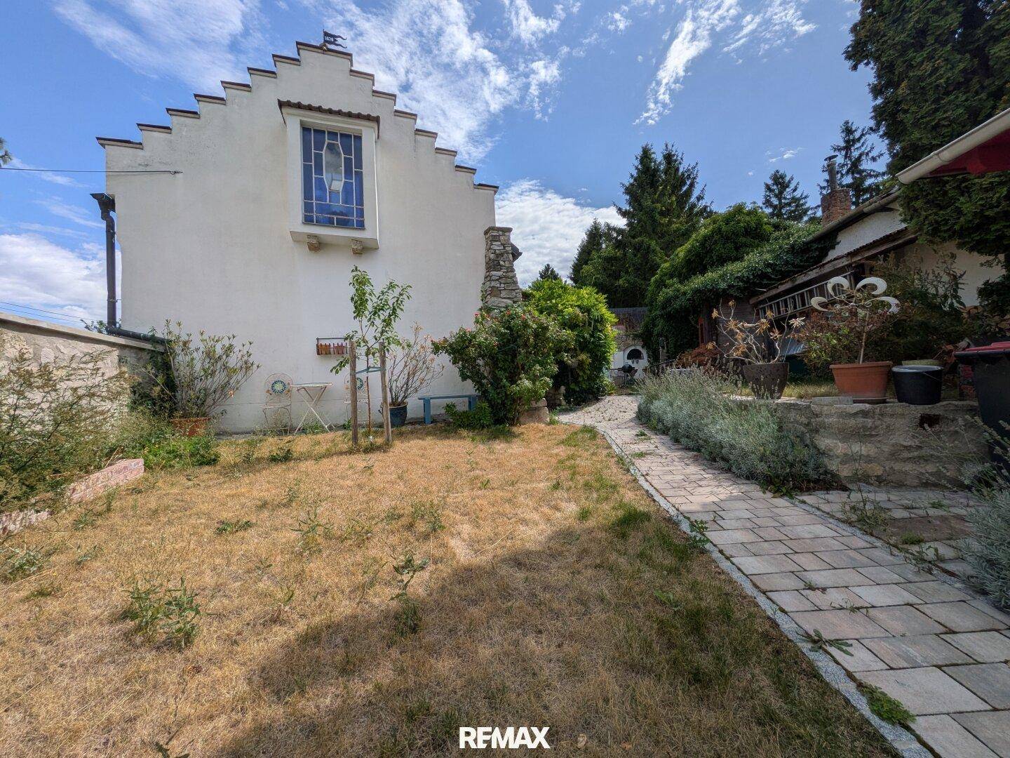 Gartenbereich hinter dem Haus mit Steinweg, trockener Rasenfläche und üppiger Vegetation.