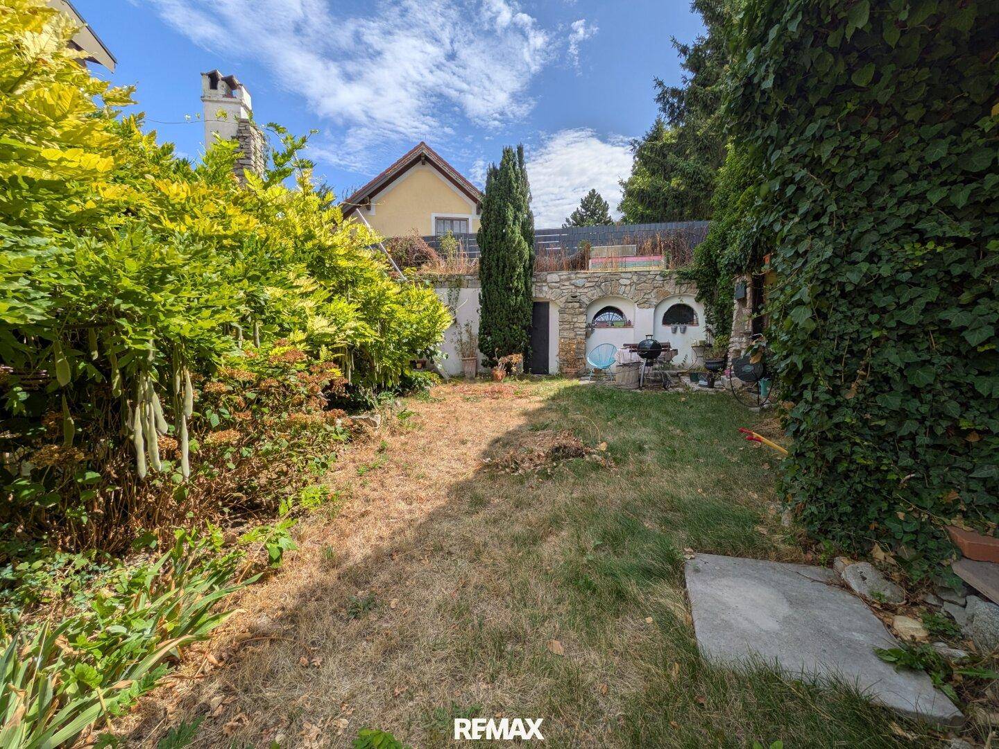 Hinterhofgarten mit Rasenfläche, verschiedenen Pflanzen und Blick auf die Rückseite des Hauses.