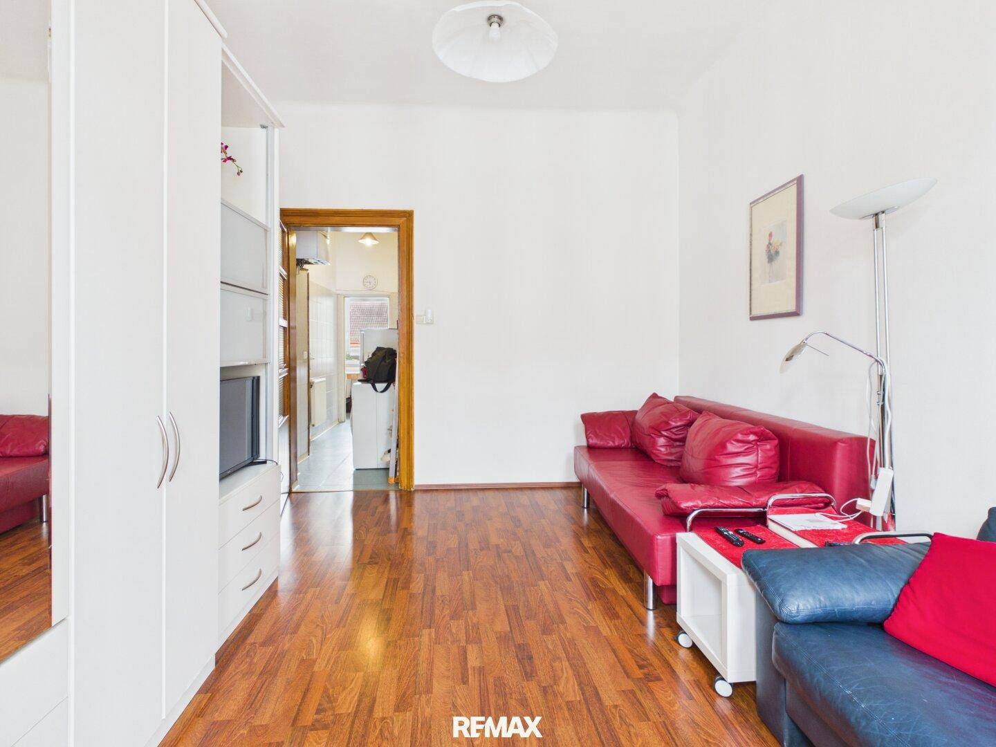 Wohnzimmer mit Holzboden, rotem Sofa und Blick durch die Tür in den angrenzenden Flur.