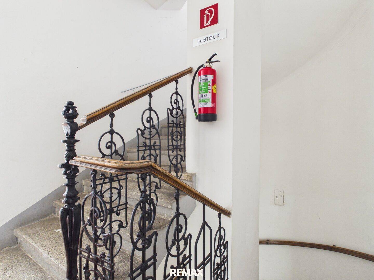 Treppenhaus mit schmiedeeisernem Geländer und einem Feuerlöscher an der Wand im dritten Stock.