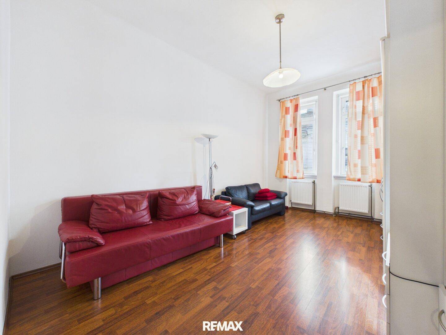 Helles Wohnzimmer mit Holzboden, zwei Fenstern und farbigen Vorhängen für eine freundliche Atmosphäre.