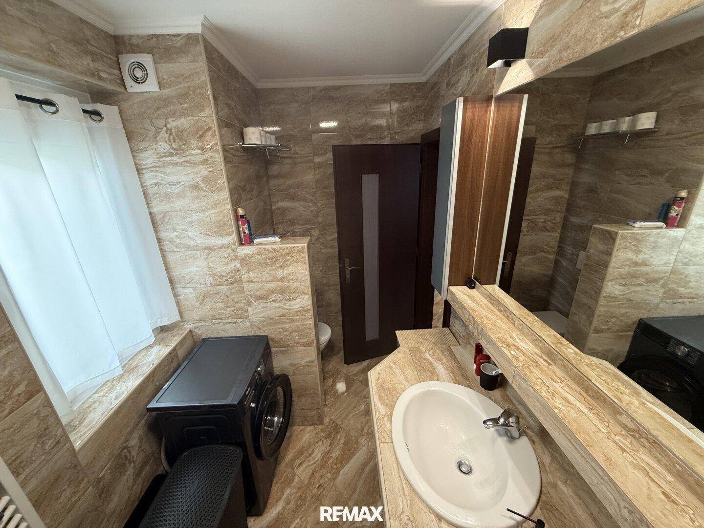 Badezimmer mit Fliesen, Einzelwaschbecken, Toilette und Fenster für Tageslicht.