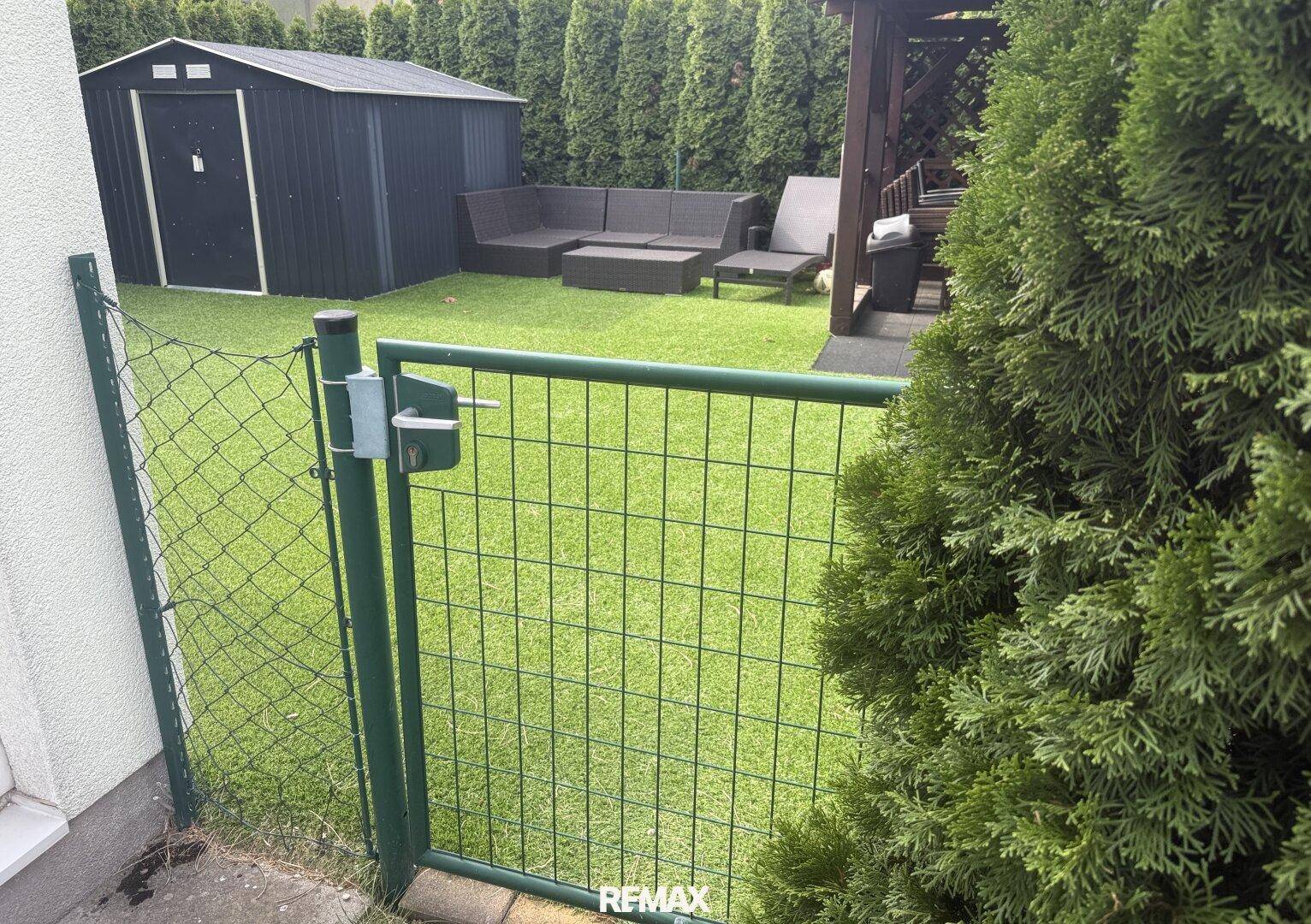 Blick auf den Gartenbereich mit Kunstrasen, Gartenhaus und Pavillon durch einen grünen Zaun.
