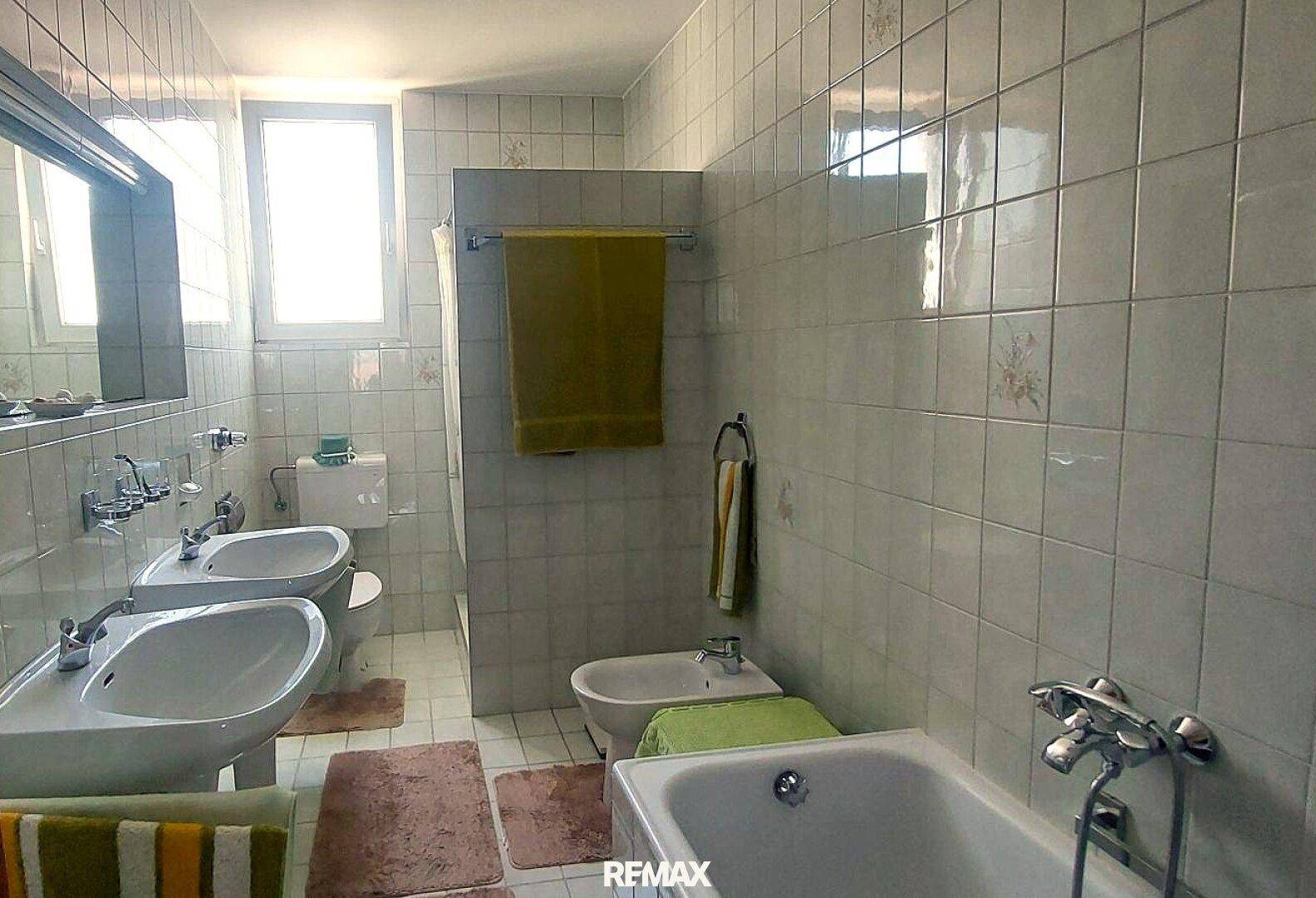 Gefliestes Badezimmer mit zwei Waschbecken, Badewanne, Bidet und einem Fenster für Tageslicht.