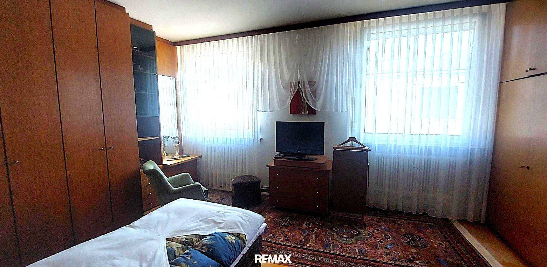 Schlafzimmer mit Einzelbett, Holzschränken, Fernseher und großen Fenstern mit Vorhängen.