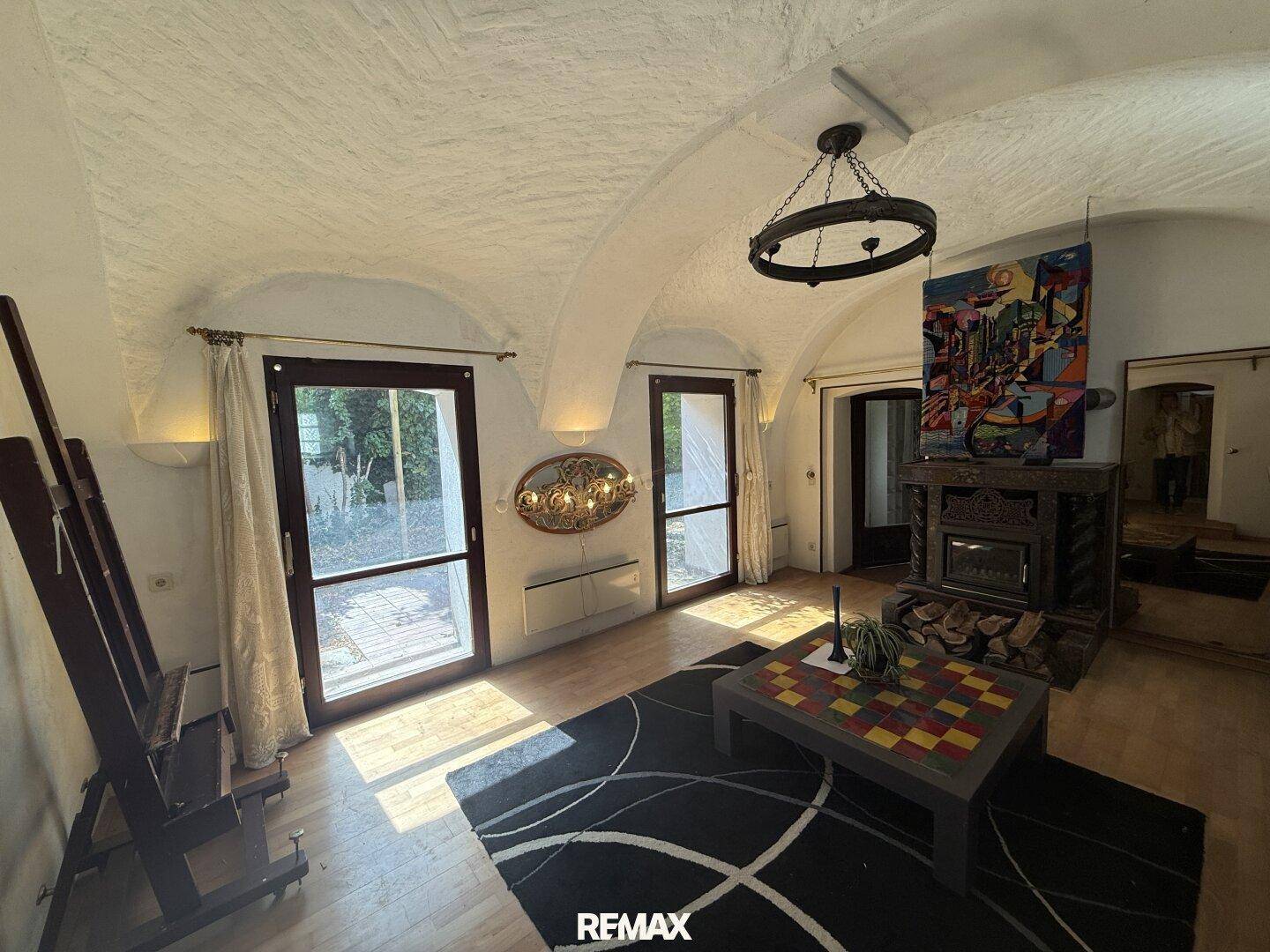 Helles Wohnzimmer mit Gewölbedecke, Kamin und Zugang zum Außenbereich durch große Türen.