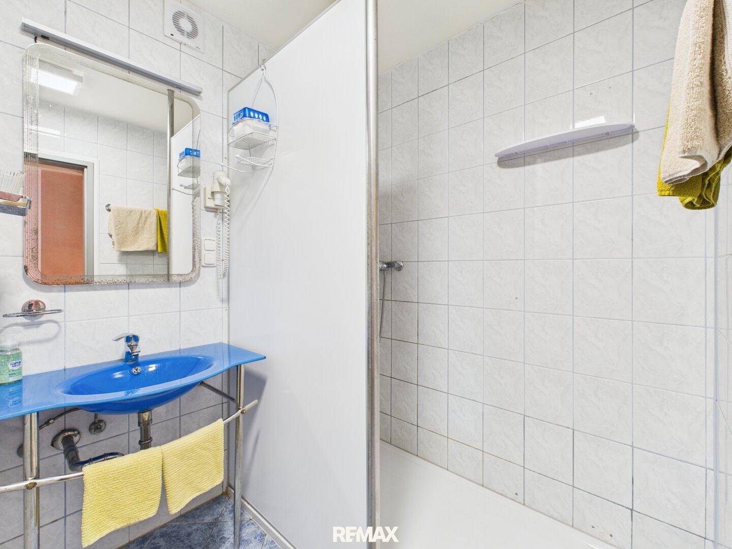 Badezimmer mit Dusche, blauem Waschbecken und weißen Fliesen, im älteren Stil gehalten.
