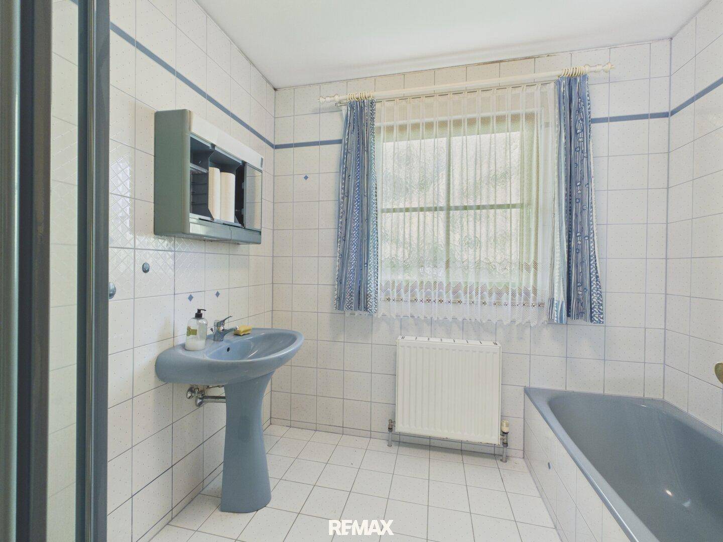 Badezimmer im 80er-Jahre-Stil mit blauen Fliesen, Waschbecken und Badewanne unter dem Fenster.
