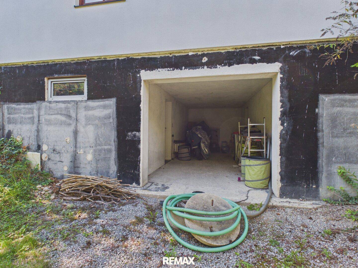 Garageneinfahrt im Untergeschoss des Hauses mit Schlauch und Holzstapel davor.