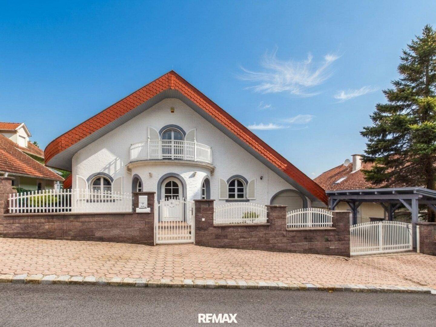 Elegante Hausfassade mit weißem Zaun, gepflasterter Auffahrt und Carport.