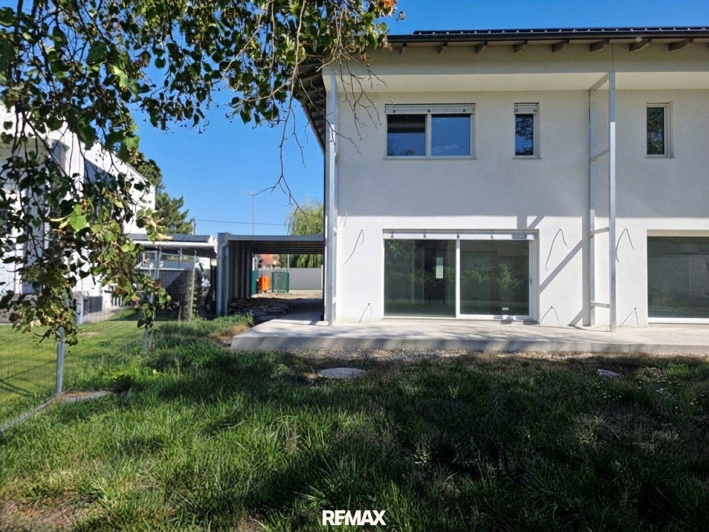 Seitenansicht des modernen Hauses mit Terrasse, Gartenbereich und angrenzendem Carport unter blauem Himmel.