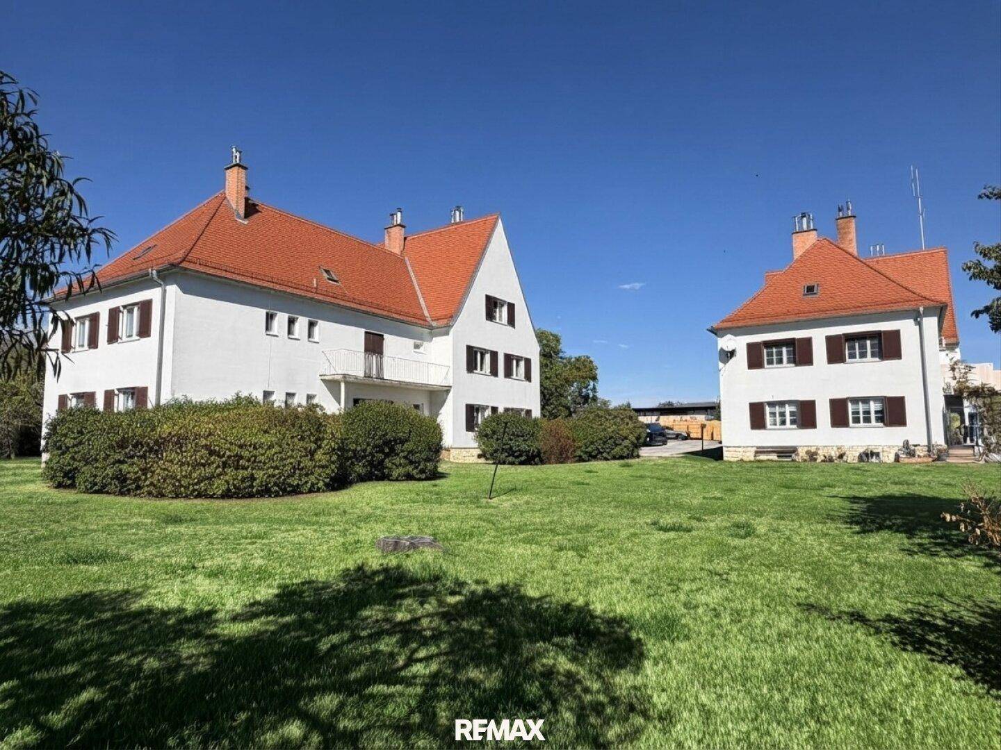 Weitläufiger Gartenbereich mit zwei traditionellen Gebäuden und gepflegten Büschen unter klarem Himmel.