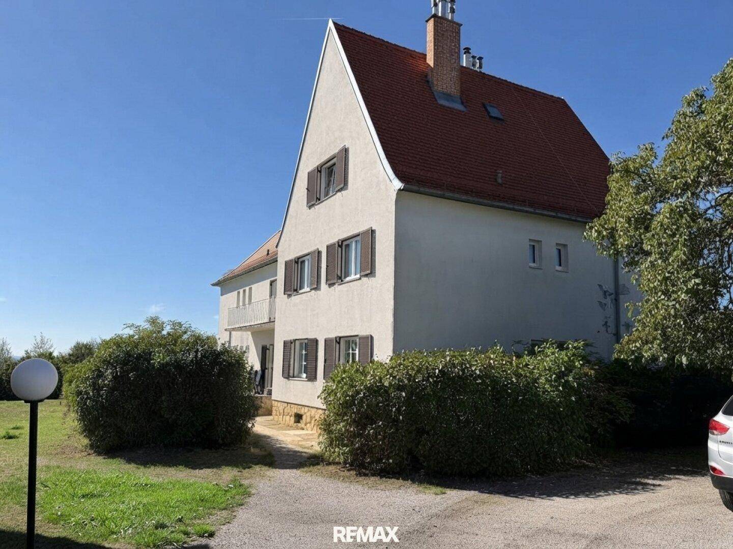 Seitenansicht eines Gebäudes mit rotem Dach, umgeben von grünen Büschen und einem Kiesweg.