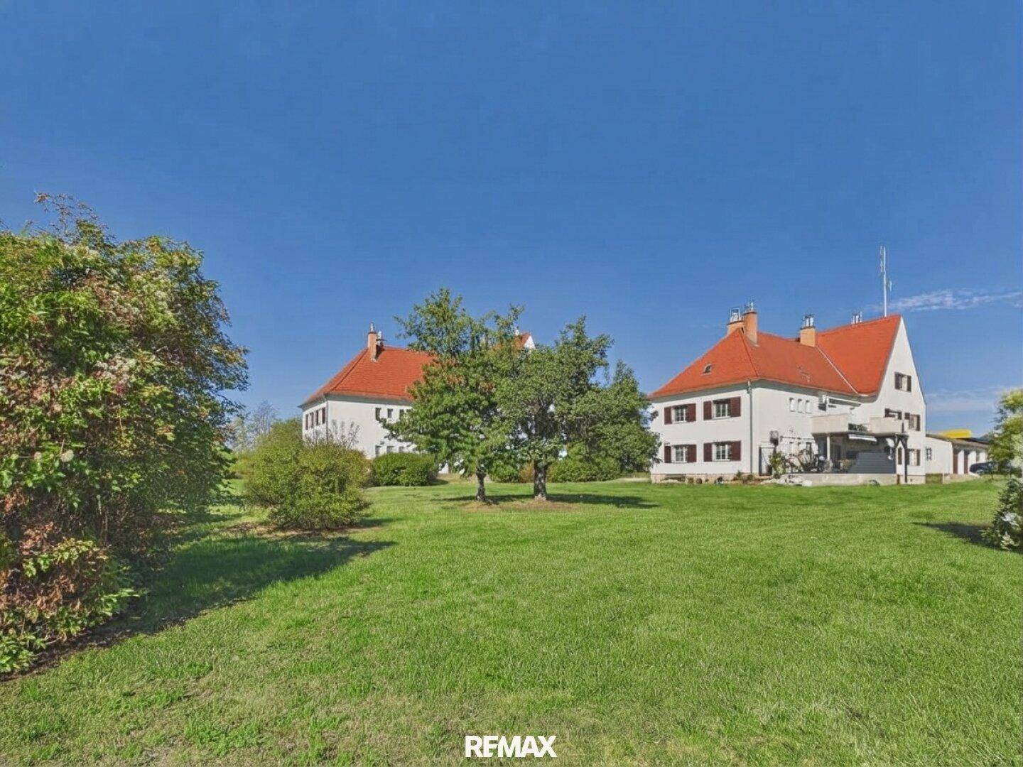 Zwei traditionelle Gebäude mit roten Dächern und einem weitläufigen grünen Rasen unter blauem Himmel.