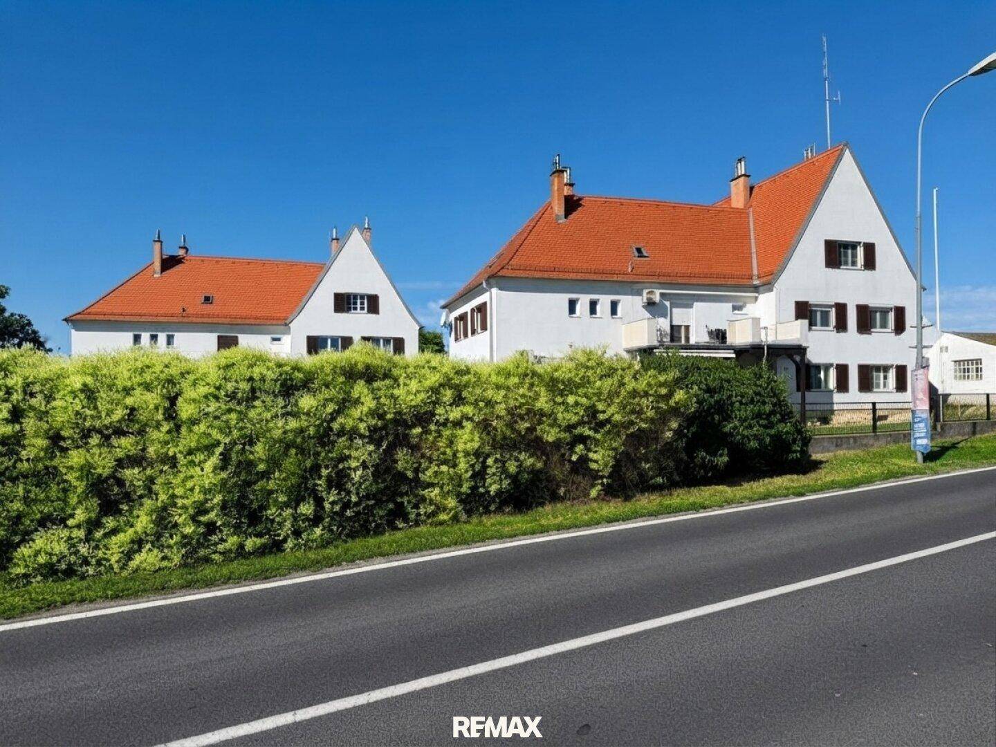 Zwei weiße Gebäude mit roten Dächern, getrennt durch eine grüne Hecke, entlang einer Straße.