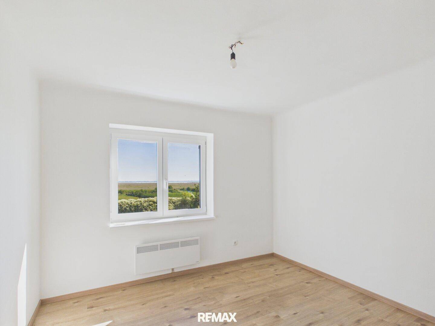 Leeres Zimmer mit weißen Wänden, hellem Holzboden und einem Fenster mit Blick ins Grüne.