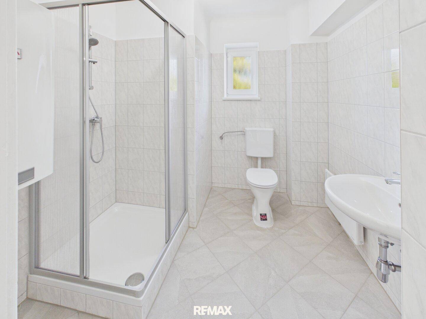 Helles Badezimmer mit weißen Fliesen, Duschkabine, Toilette, Waschbecken und einem Fenster.
