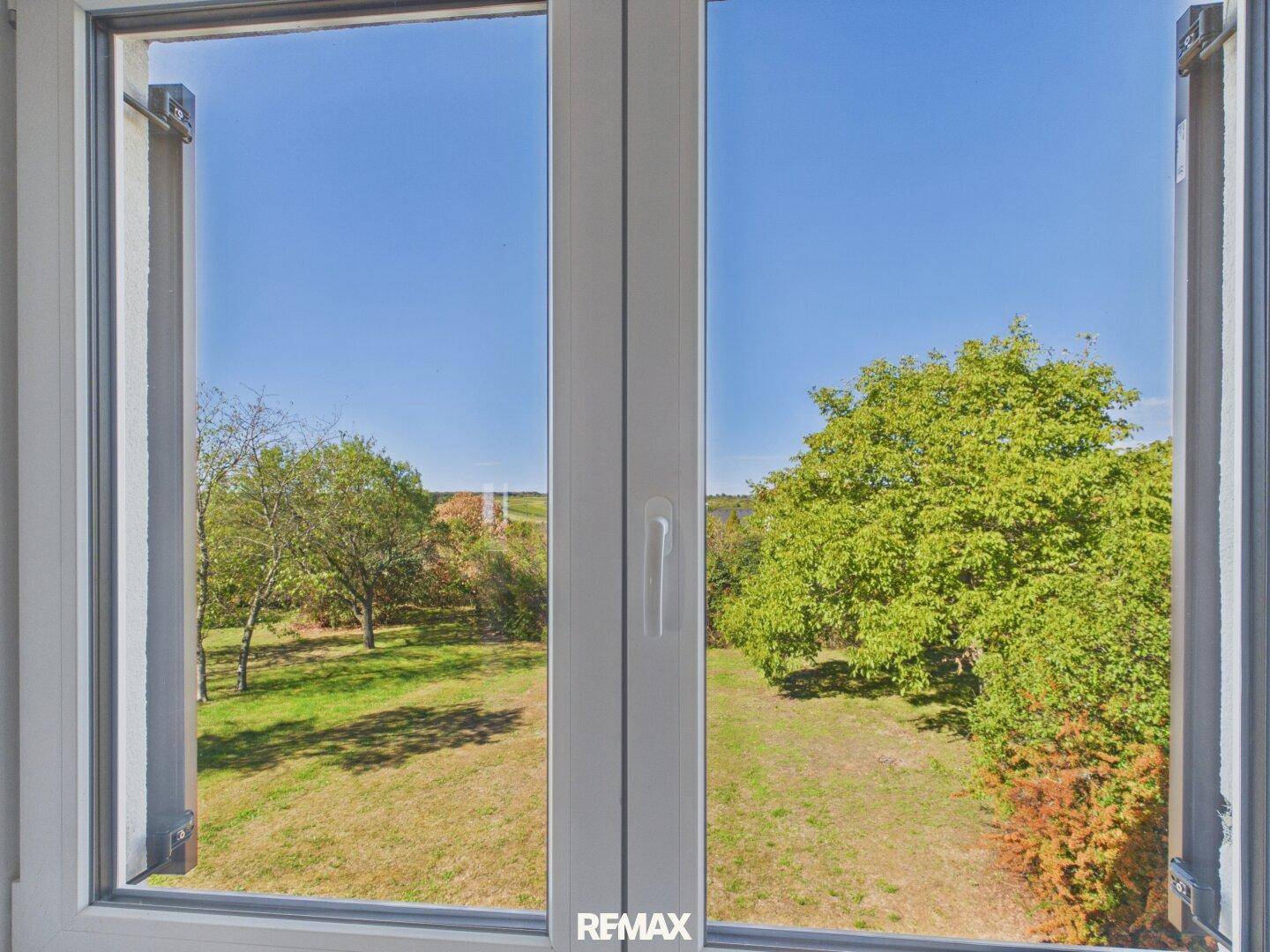 Fenster mit Blick auf einen grünen Garten und Bäume unter blauem Himmel.