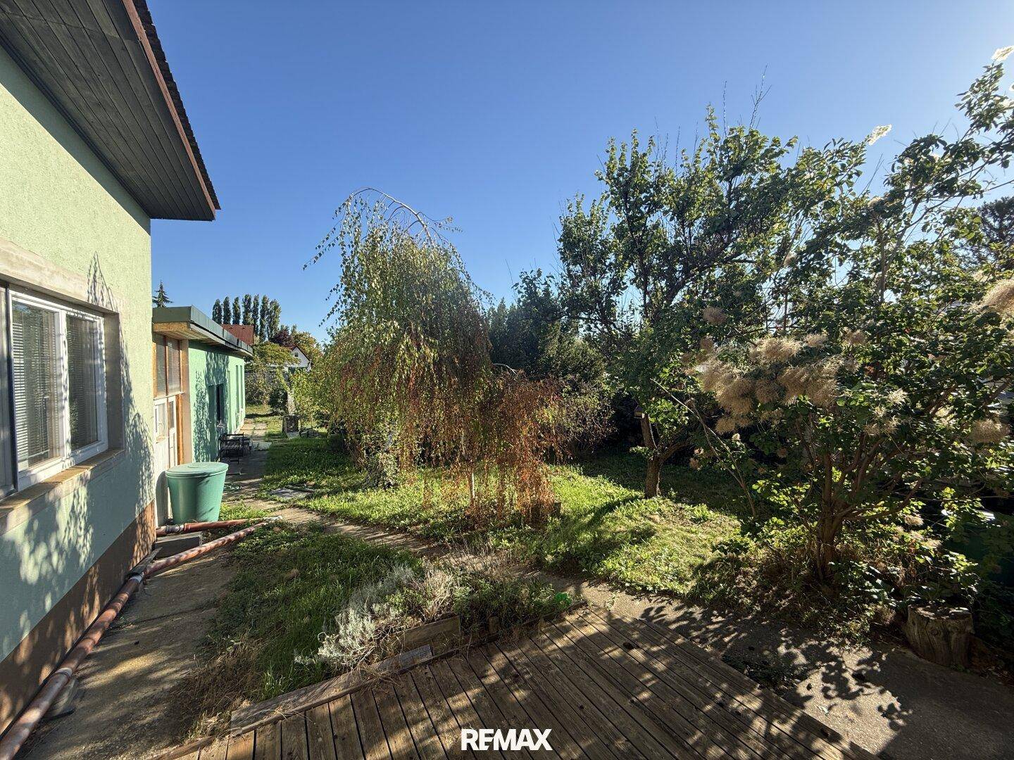 Sonniger Garten mit altem Baumbestand und einer kleinen Holzterrasse am Haus.