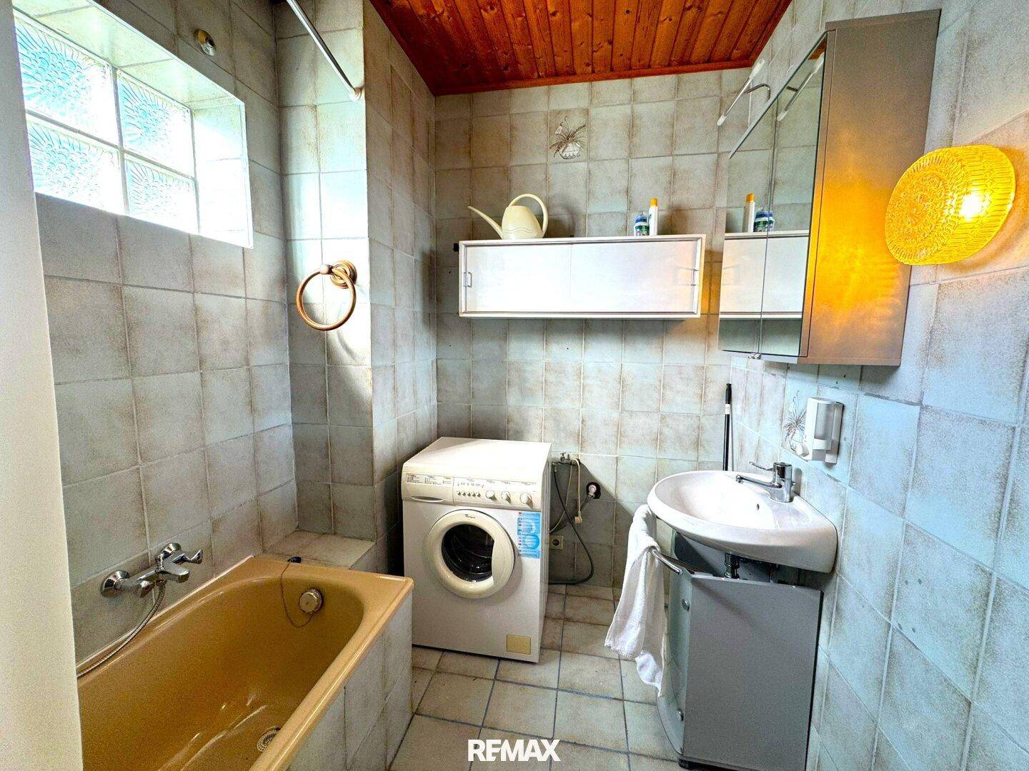 Badezimmer mit Badewanne, Waschmaschine und Holzdecke, ausgestattet mit Fliesen.
