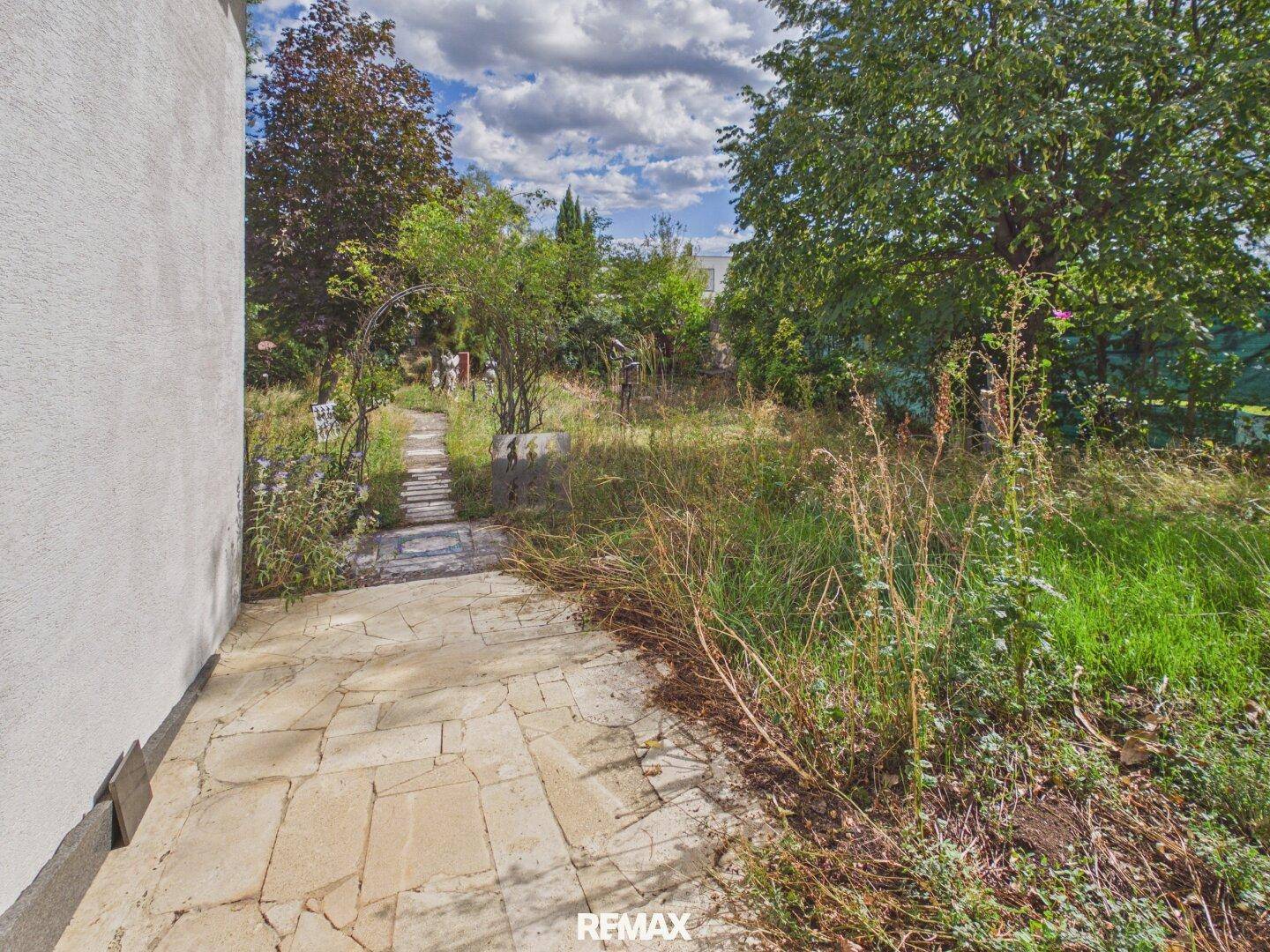 Steinplattenweg führt durch den verwilderten Garten mit üppiger Vegetation.