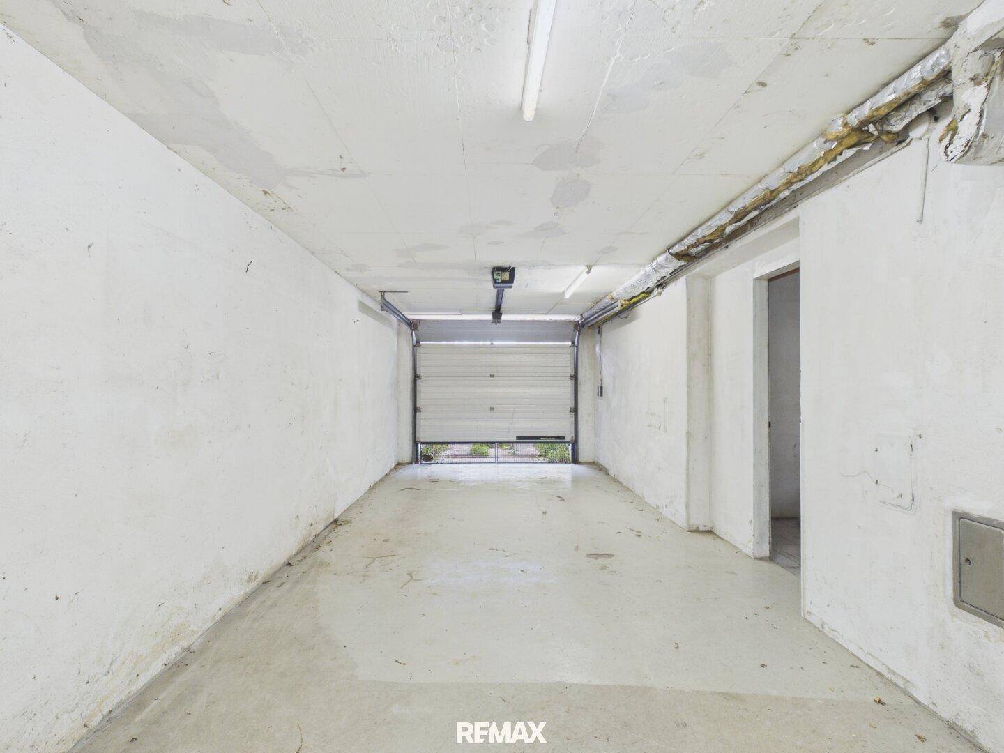 Leere Garage mit Betonboden, weißen Wänden und einem geöffneten Rolltor.