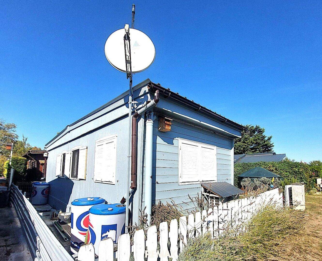 Blaues Holzhaus mit Satellitenschüssel, Regentonnen und weißem Zaun.