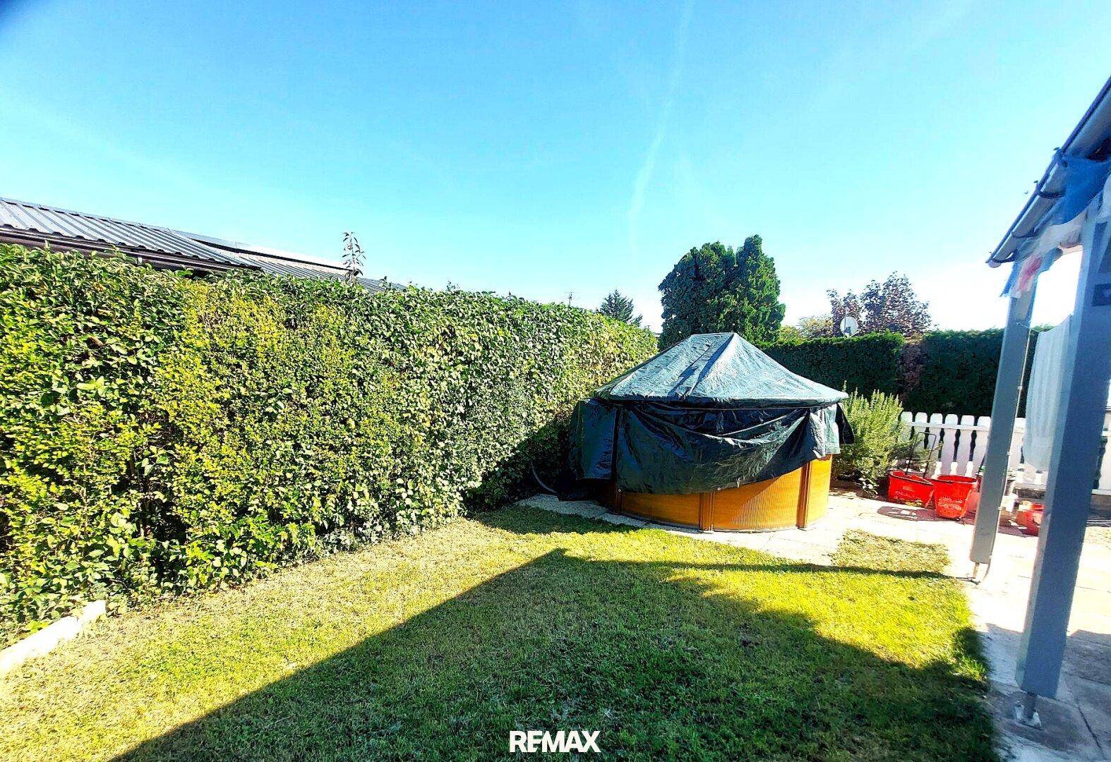 Grüner Garten mit Rasenfläche, hoher Hecke und einem überdachten Whirlpool oder Pool.
