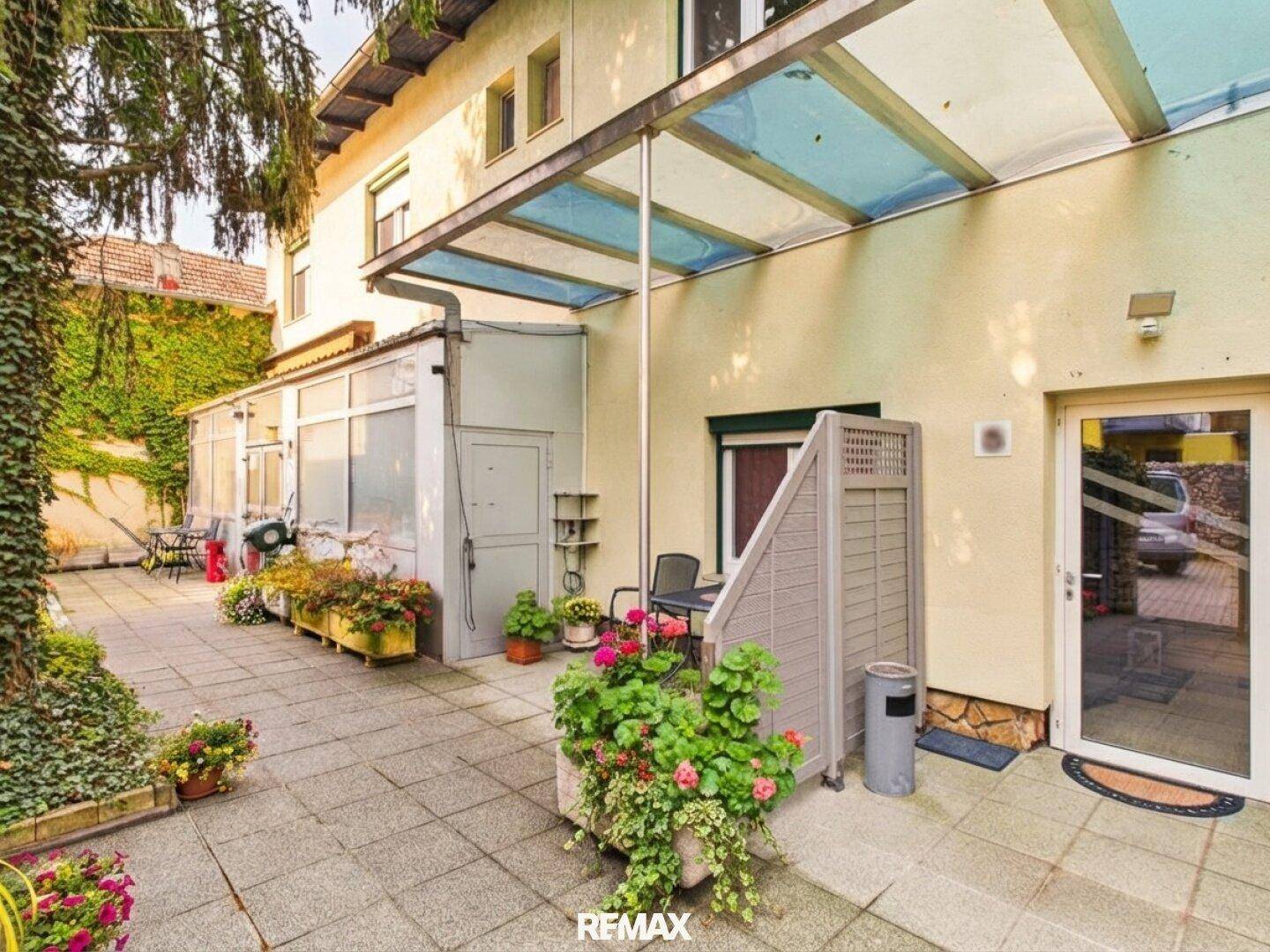 Außenbereich mit überdachter Terrasse, Pflanzen und Zugang zum Gebäude.