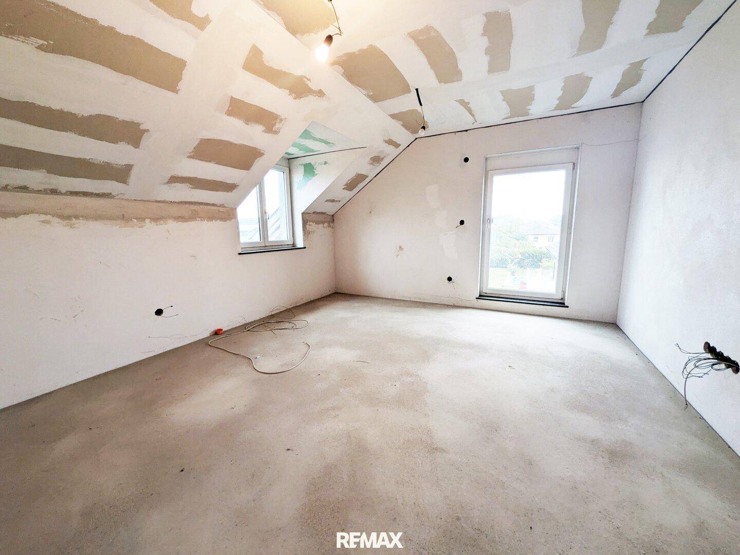 Dachgeschosszimmer im Rohbau mit schrägen Wänden und Fenster, bereit für den Ausbau.