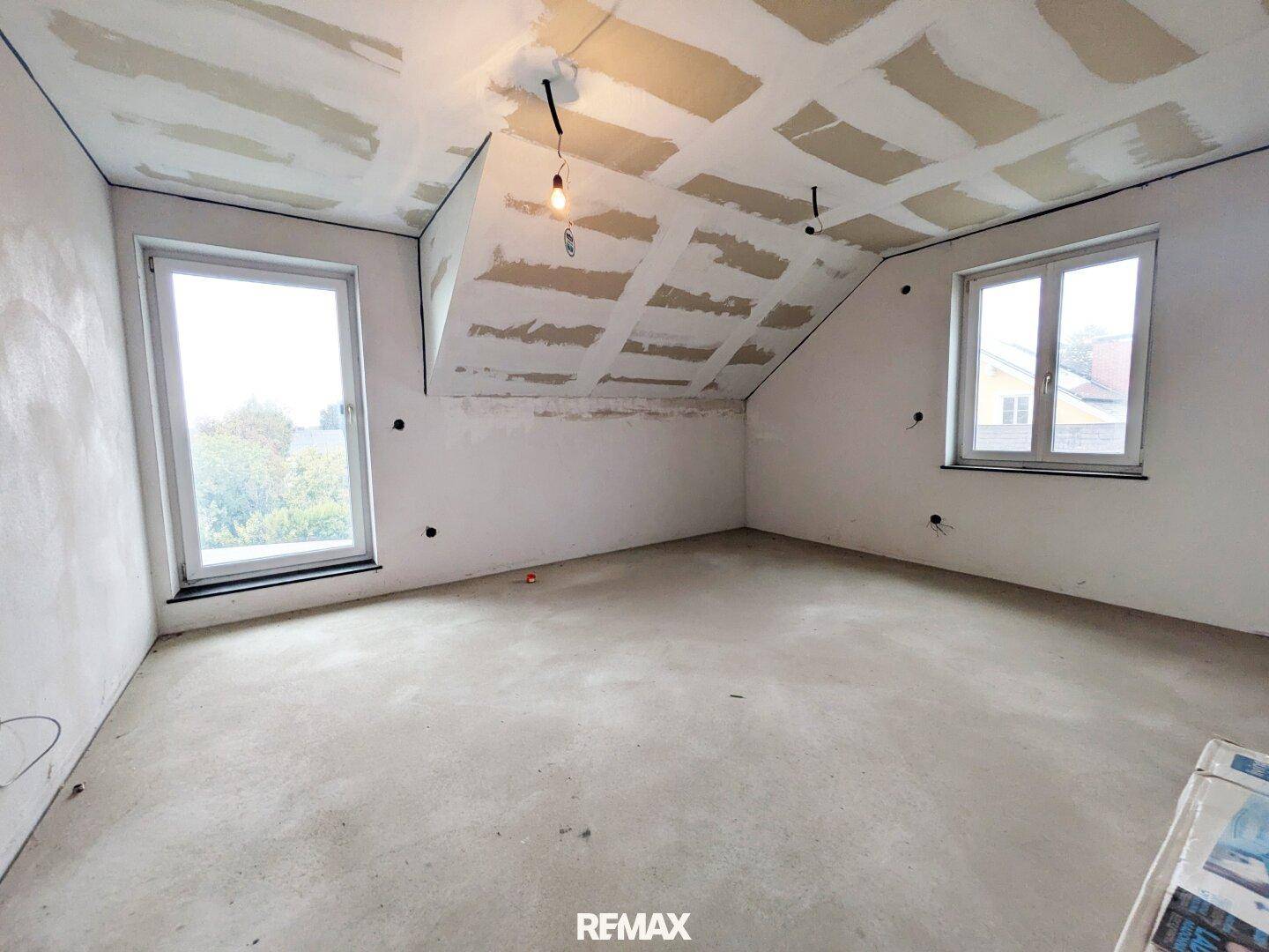 Dachgeschossraum im Rohbau mit schrägen Wänden und zwei Fenstern, viel Potenzial.