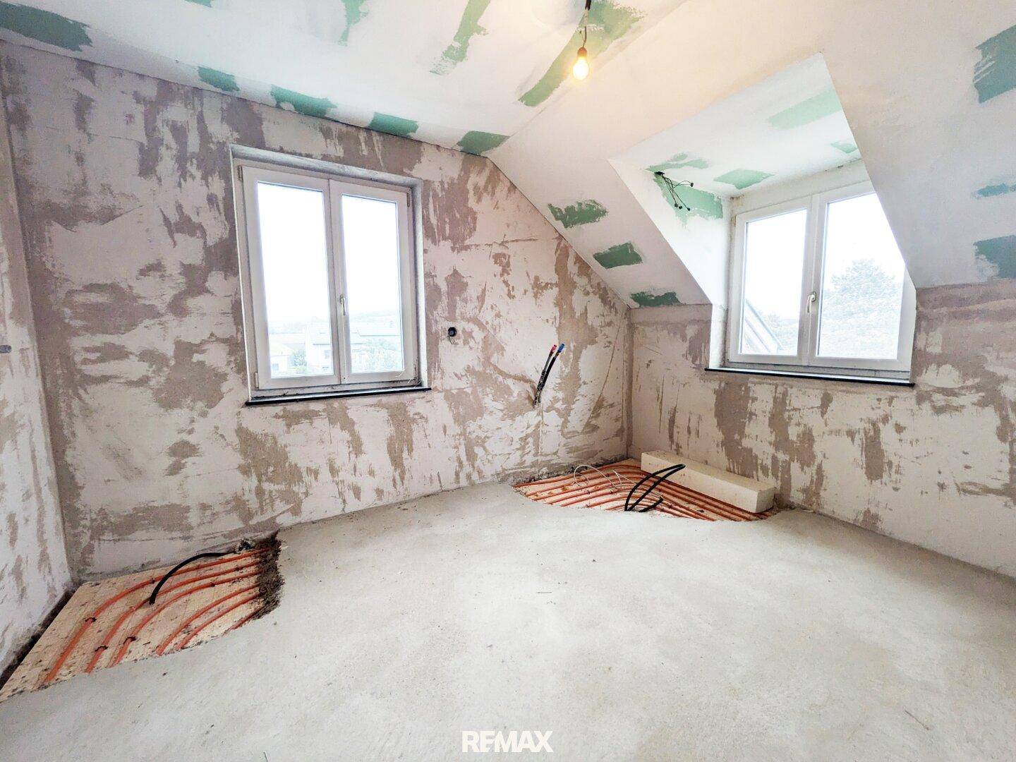 Dachgeschosszimmer im Rohbau mit sichtbarer Fußbodenheizung und zwei Fenstern.