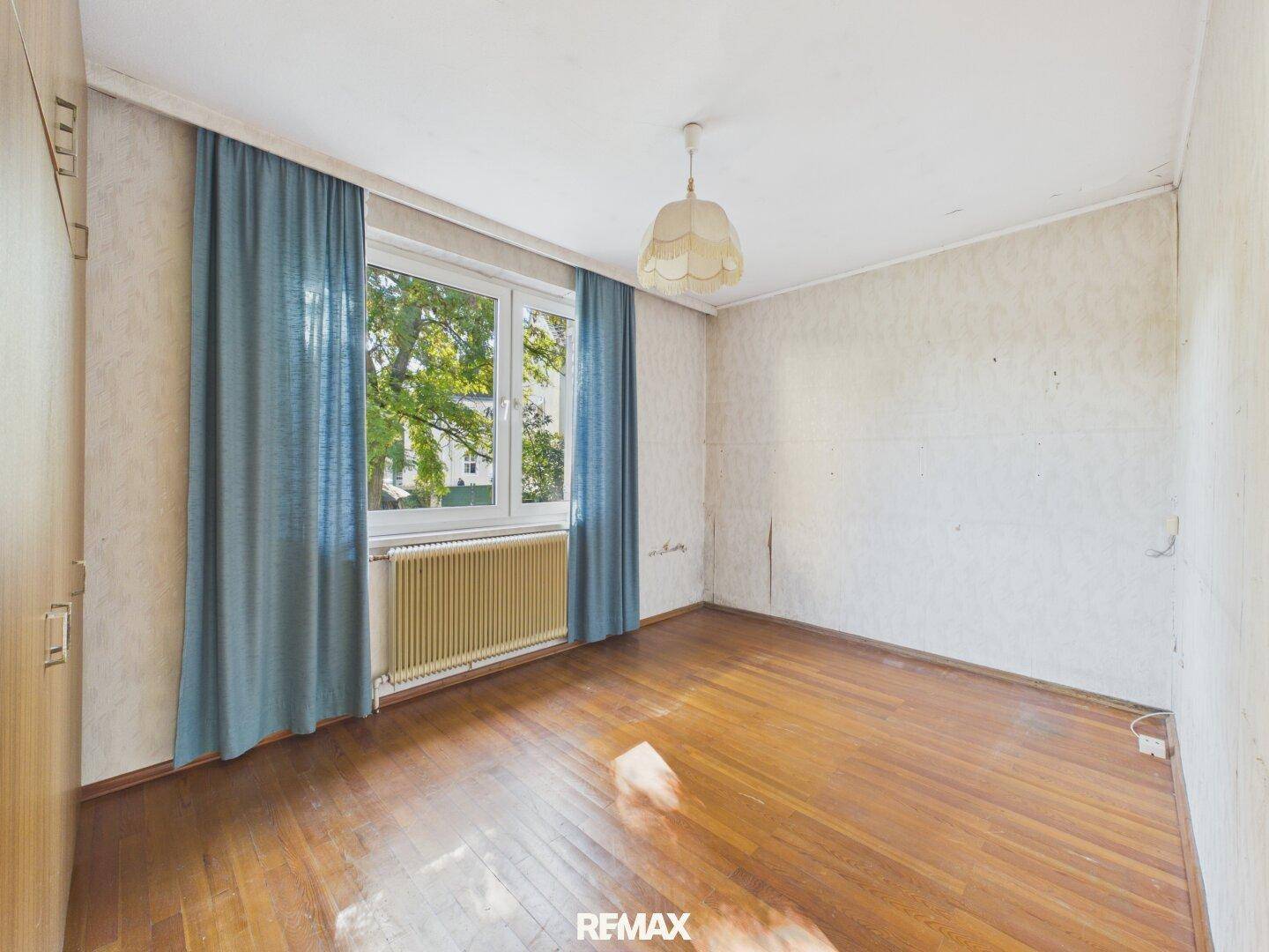 Schlafzimmer mit Holzparkettboden, großem Fenster, blauen Vorhängen und Heizkörper.