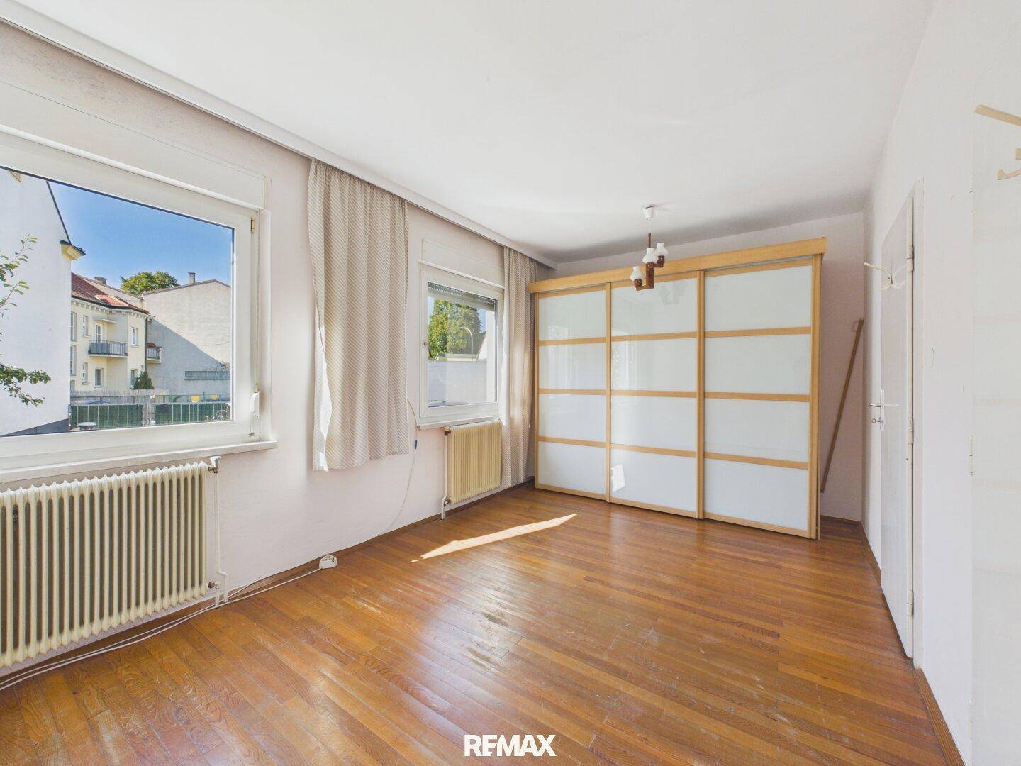 Helles Schlafzimmer mit Holzparkettboden, Schiebetürenschrank und zwei Fenstern.