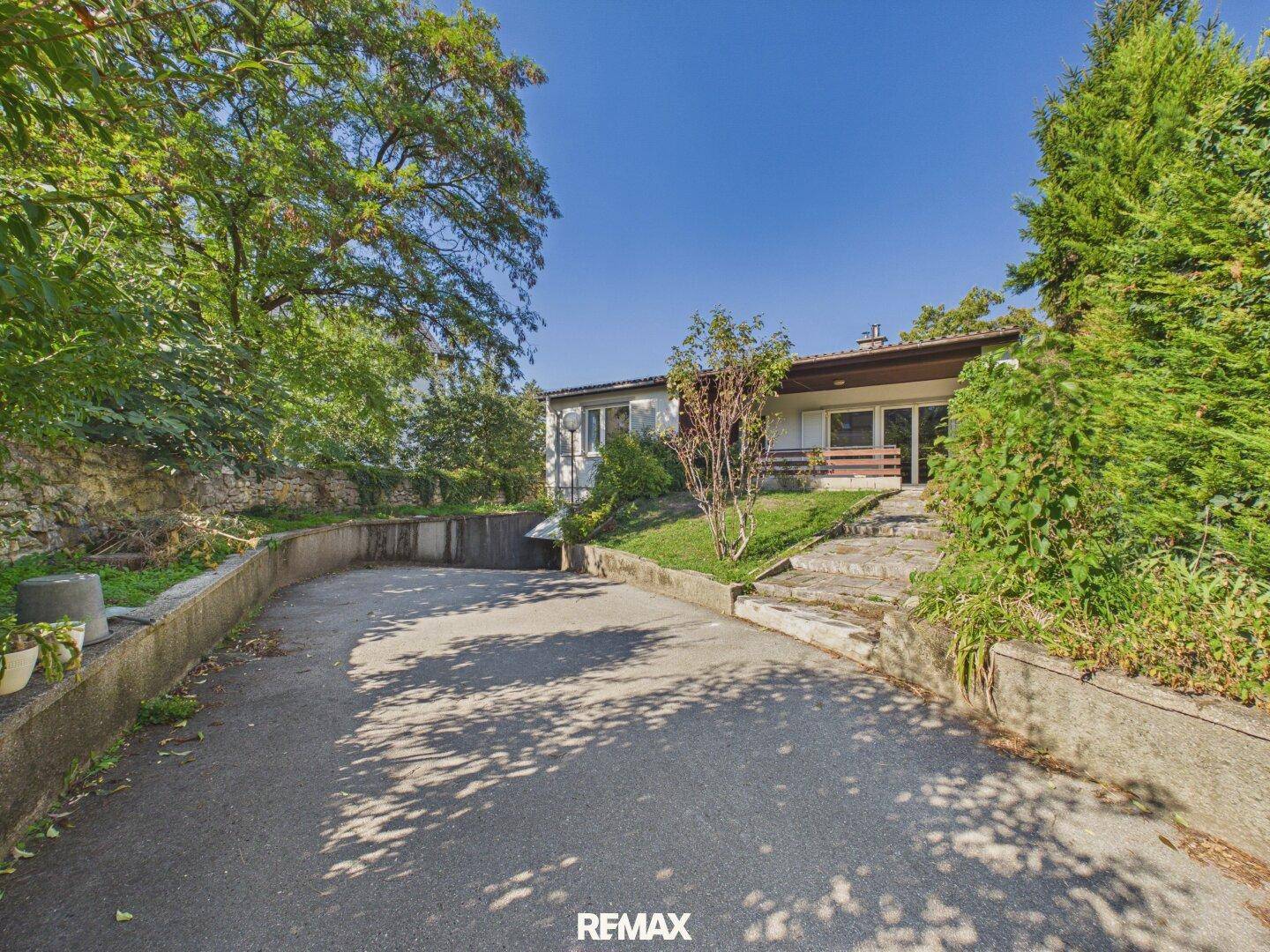 Einfahrt zum Haus mit Zugang zur Garage und umgebenden Bäumen und Sträuchern.