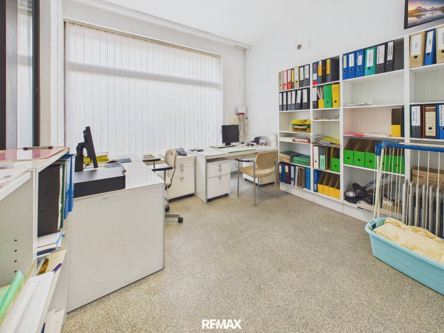Geräumiges Büro mit mehreren Arbeitsplätzen, einem großen Fenster und einem Regal für Aktenordner.
