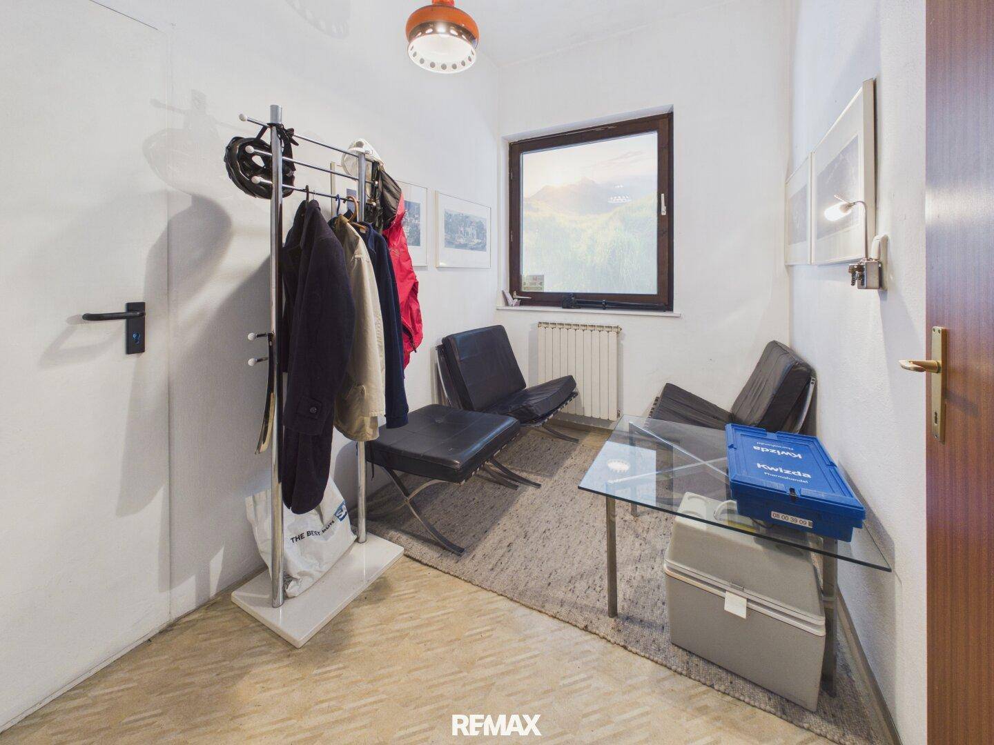 Kleiner Wartebereich oder Flur mit Garderobe, zwei Sesseln und einem Glastisch, beleuchtet durch ein Fenster.