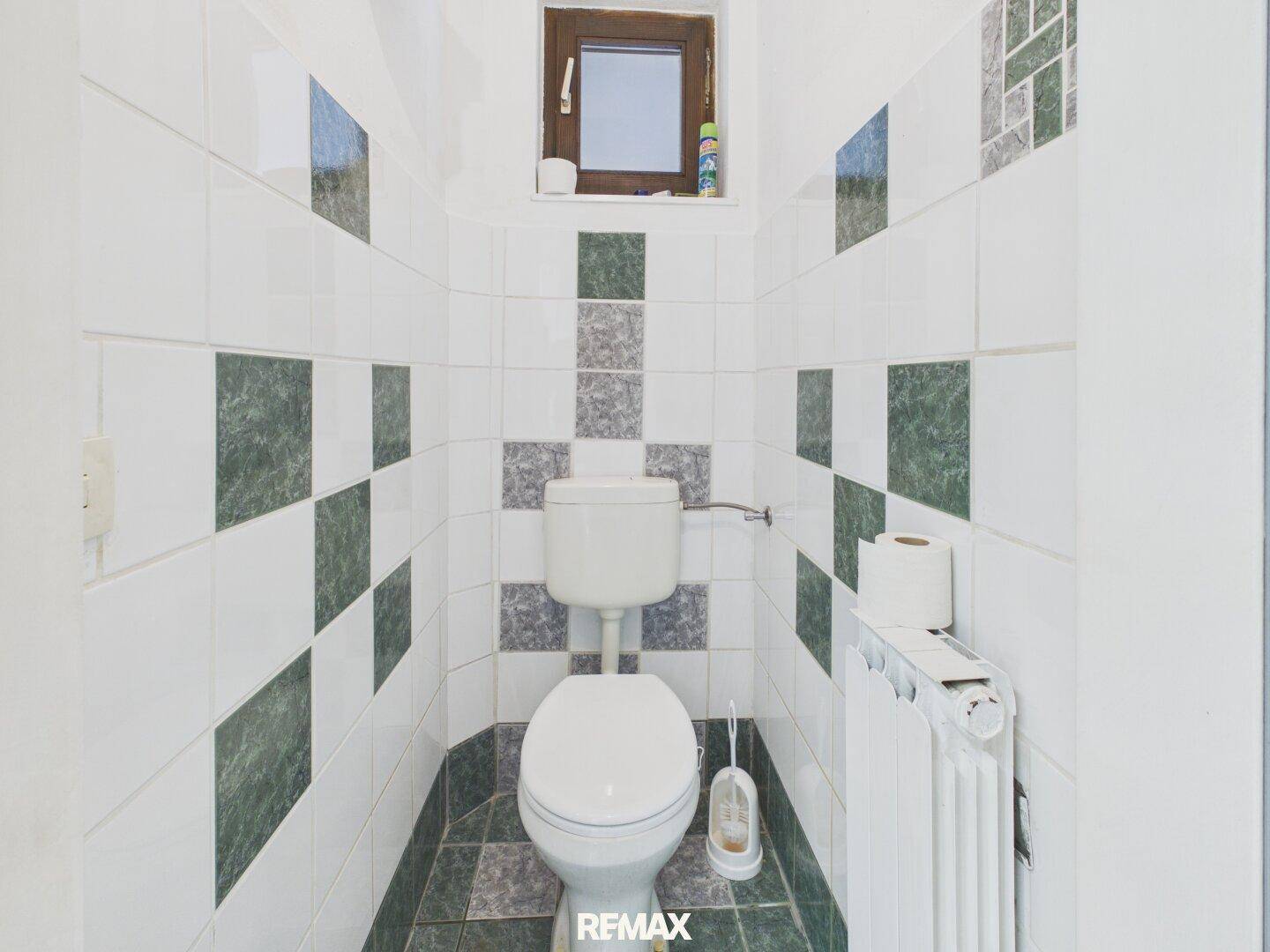 Toilettenraum mit WC, Heizkörper und gemusterten Fliesen in Grün- und Grautönen, ergänzt durch ein kleines Fenster.